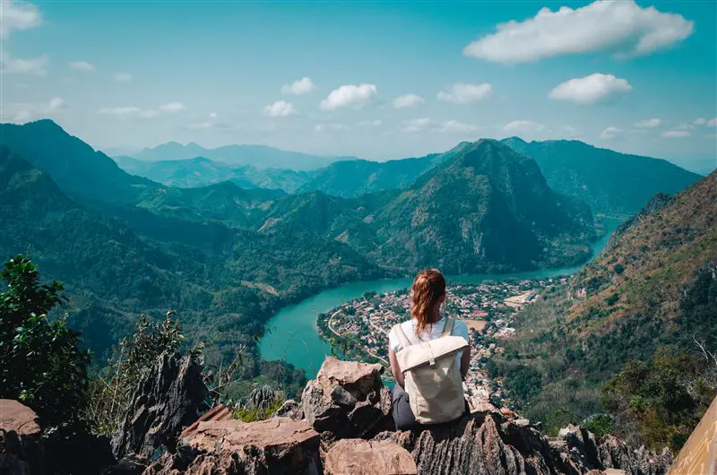 A woman wearing a backpack sits on a rock with a view of a s-shaped river and town from Pha Daeng Peak in Nong Khiaw, Laos