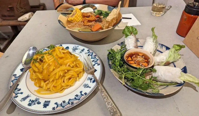 A table with three plates of vegan food - a plate of pumpkin spaghetti, four fresh spring rolls, and a vegetarian noodle soup - at Thong Dong Vegan Kitchen in Sapa, Vietnam