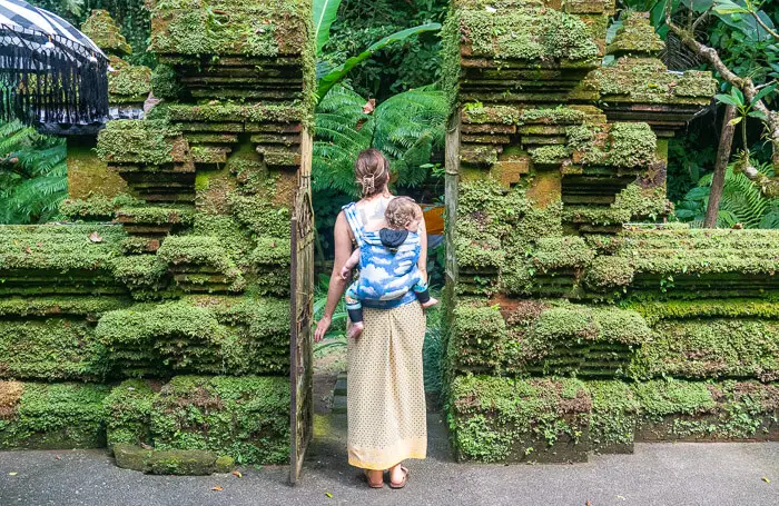 A woman carrying a baby on her back and walking through a moss-covered temple gate in Bali