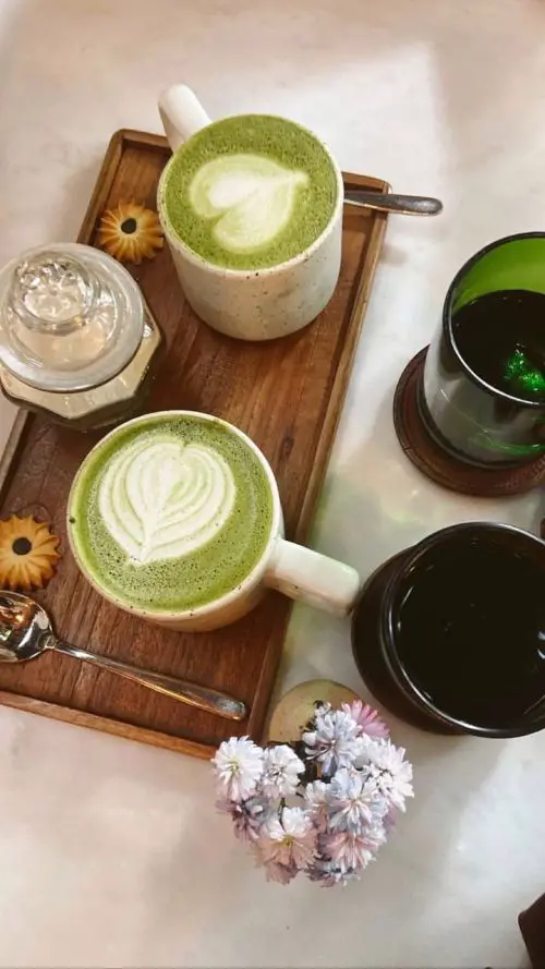 Two mugs of matcha latte with leaf art on a white table with flowers