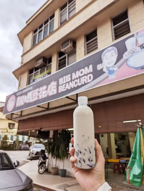 A hand holding up a plastic bottle of fresh soy milk with grass jelly in front of Big Mom Beancurd storefront in Ipoh, Malaysia