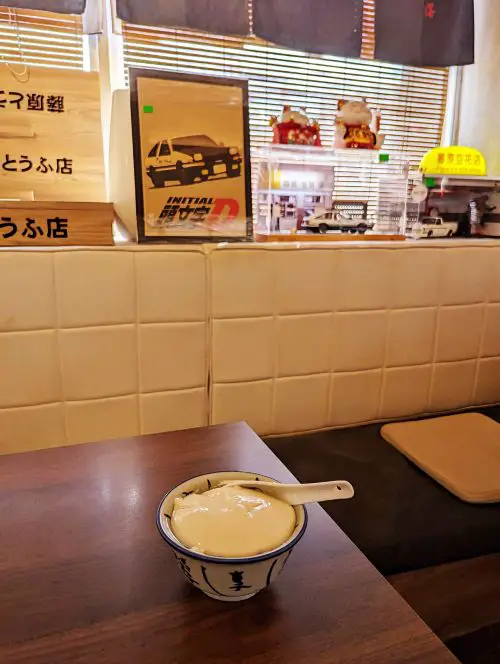 A bowl of tau fu fa tofu dessert with Initial D posters and trinkets inside Ipoh's Fujiwara Tofa Shop