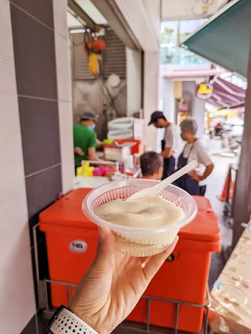A hand holding a plastic bowl of tau fu fa tofu dessert at Funny Mountain Soya Beancurd in Ipoh