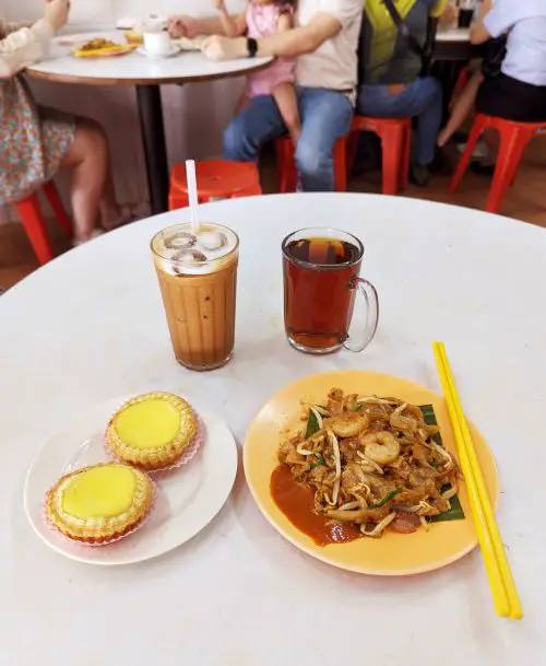 A table with a plate of egg tarts, char kway teow fried noodles, iced coffee, and hot tea at Ipoh's Kedai Manakan Nam Heong shop