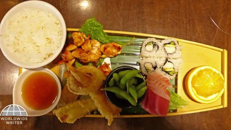 A wooden boat of tempura, chicken teriyaki, sashimi, edamame, and California rolls and a side of rice at a restaurant in Tokyo, Japan