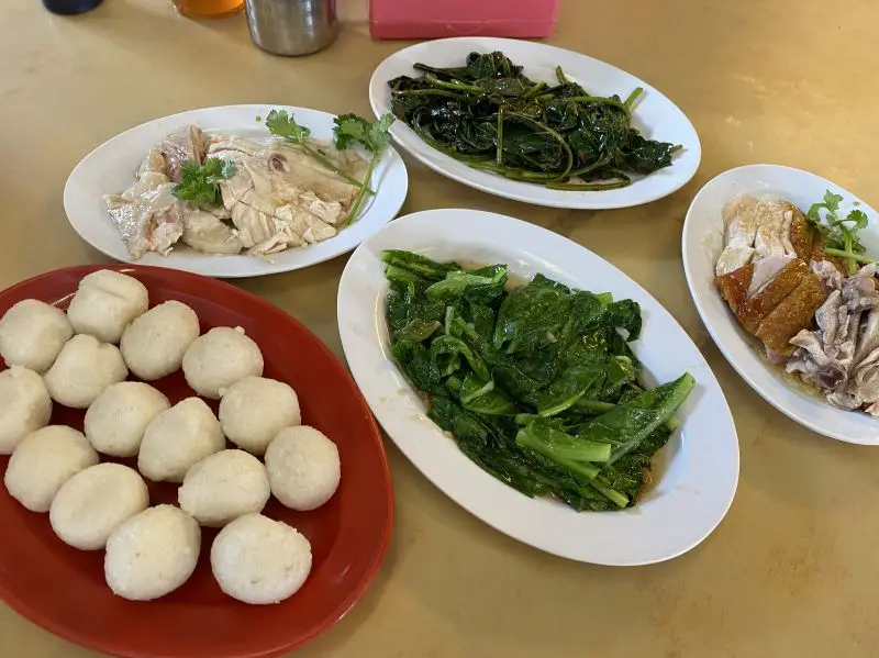 A table with five plates of Malaysian food including two plates of green stir fried vegetables, two plates of chicken, and one plate of chicken rice balls at Ee Ji Ban Chicken Rice Ball in Melaka, Malaysia