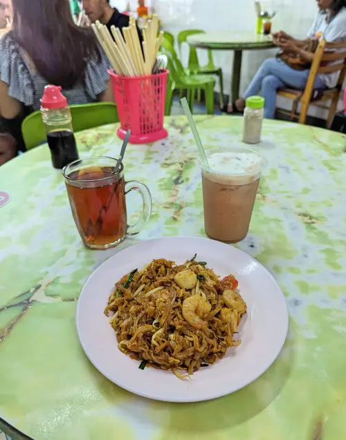 A green and white table with an iced coffee, a hot tea, and a plate of char kway teow, fried noodles with shrimp and fish balls at Bee Hwa Cafe in Penang