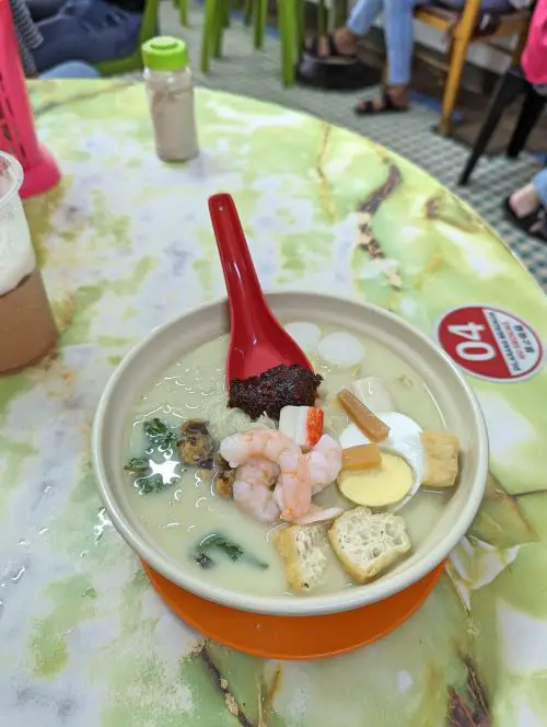 A bowl of halal white curry noodles with shrimp, egg, and fish cakes and a spoon with chili paste at Bee Hwa Cafe in Penang, Malaysia