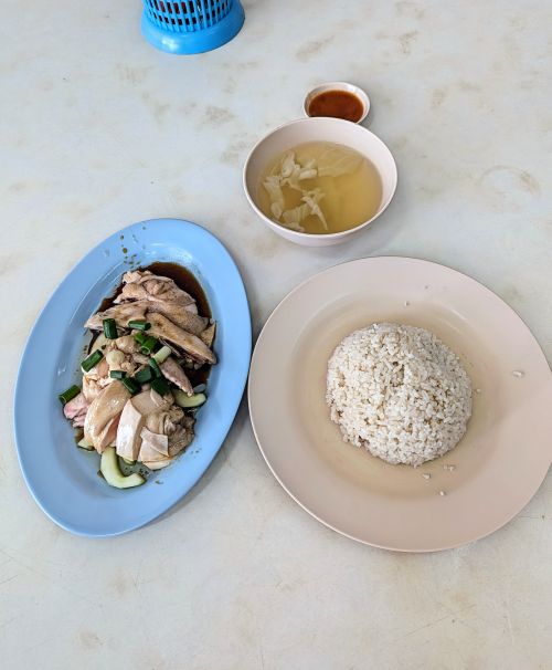 A light blue plate of chicken with a light pink plate of chicken flavored rice, and a bowl of soup at Gow Thew Chik Hainan Chicken Rice in Penang