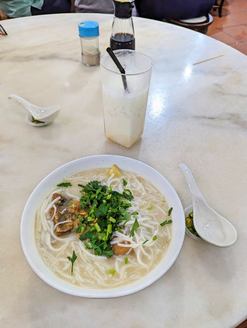 A bowl of fish head noodle soup with rice noodles and a cup of iced almond milk at Hon Kei Food Corner in Penang