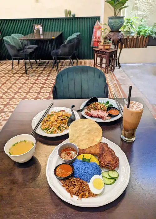 A table at Hwa Ting of a bowl of soup, an iced beverage, and three plates of Malaysian food, a nasi lemak with fried chicken and blue and yellow rice, a plate of noodles with roasted pork, and a plate of fried noodles