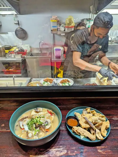 A bowl of clam noodle soup and a plate of fried fish skin on a counter table and a person working in the kitchen in the background at Penang's Ichi Tong