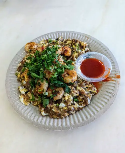 A paper plate of shrimp omelette with a side of chili sauce from Penang Road Famous Laksa