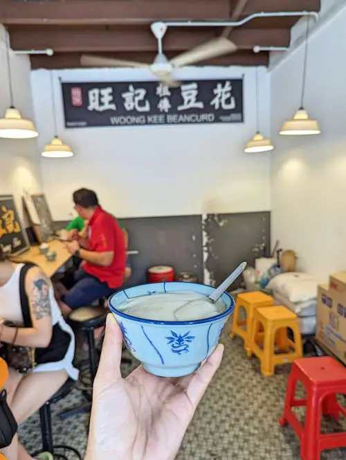 A hand holding a blue and white floral bowl with soft tofu pudding inside at Woong Kee Beancurd in Penang