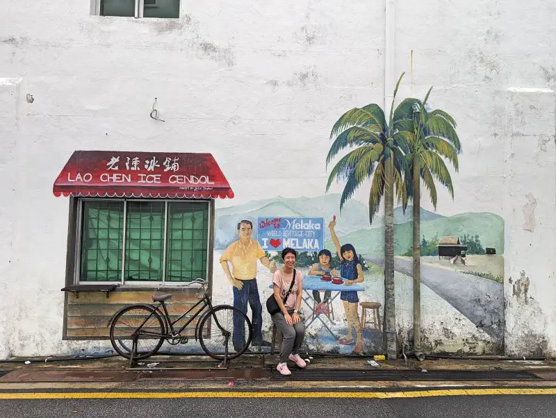 Jackie Szeto, Life Of Doing, sits on the stool in front of the Lao Chen Ice Cendol street art in Melaka, Malaysia