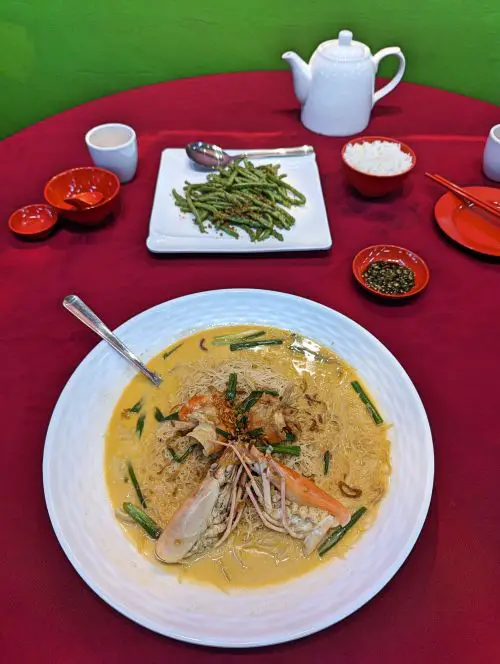 A plate of cheesy prawn mee hoon noodles and a plate of green beans on a burgundy colored table cloth at Restoran Tong Sheng in Melaka, Malaysia