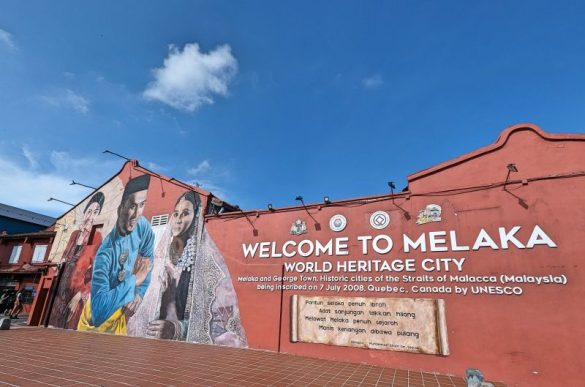 A red brick colored wall with a Welcome to Melaka World Heritage City sign and murals of two Malaysian woman and one Malaysian man