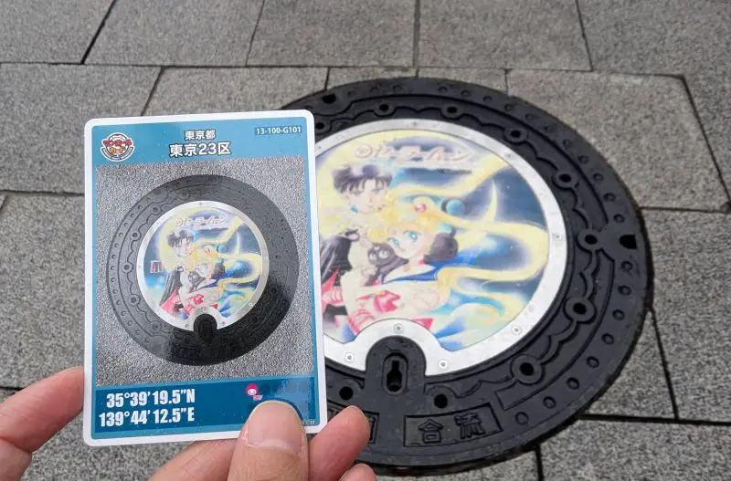 A hand from Life Of Doing holds the manhole card that matches the Sailor Moon, Tuxedo Mask, and Luna manhole cover in Tokyo, Japan