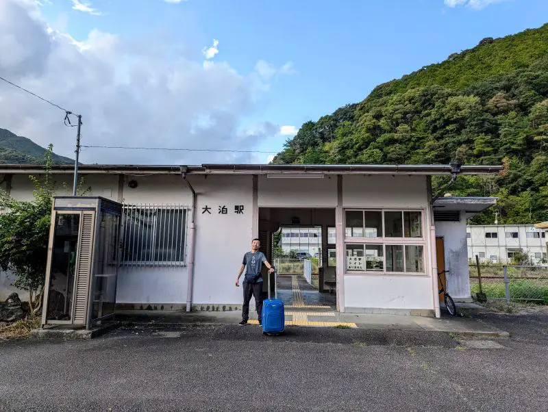 Justin Huynh, Life Of Doing, stands with a blue rolling luggage in front of Odomari Station in Kumano, Japan