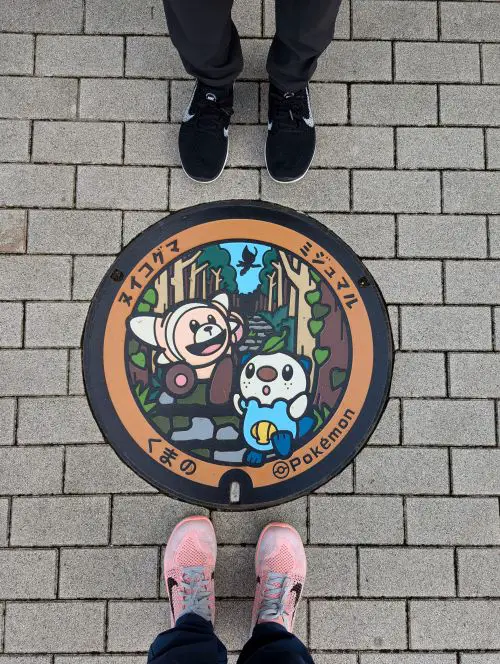 Two sets of feet from Life Of Doing stand around the Pokemon manhole cover lid