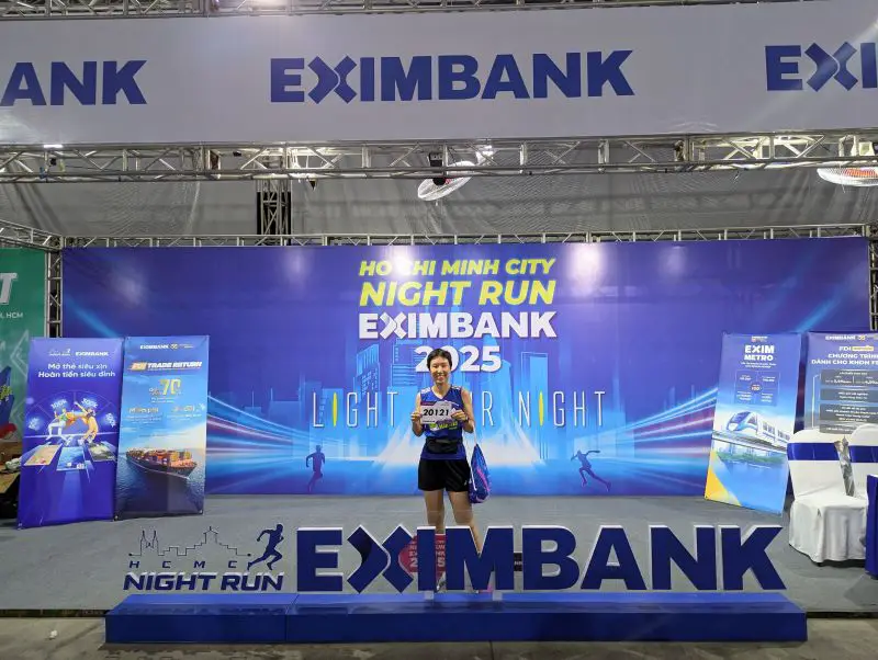 Jackie Szeto, Life Of Doing, holds her racing bib and stands behind the HCMC Night Run Eximbank signage at the Expo