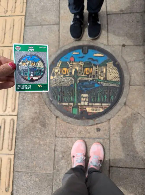 Jackie Szeto, Life Of Doing, holds the Otaru manhole card and stands near the matching Otaru Canal manhole cover