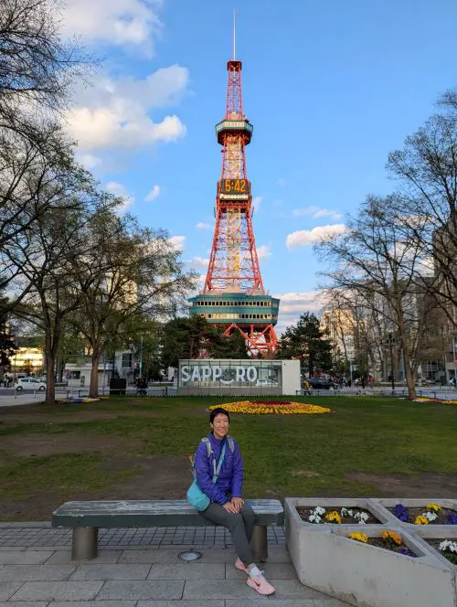 Jackie Szeto, Life Of Doing, sits on a bench in front of the Sapporo Tower in Sapporo, Hokkaido, Japan