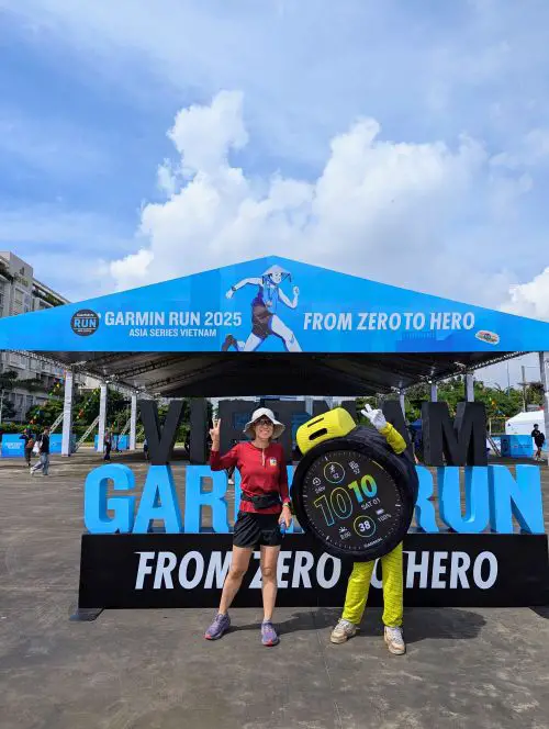 Jackie Szeto, Life Of Doing, holds a peace sign and stands next to the Garmin Forerunner 965 mascot at the Garmin Run Vietnam Expo