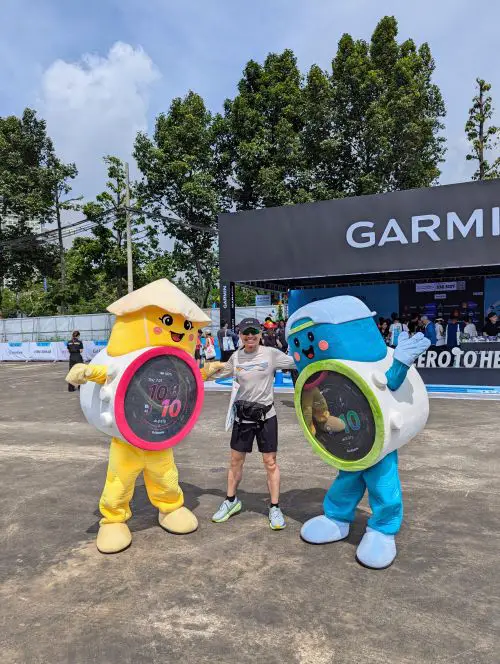 Justin Huynh, Life Of Doing, stands in between the Garmin Forerunner 970 and Forerunner 570 mascots at the Garmin Run Vietnam Expo