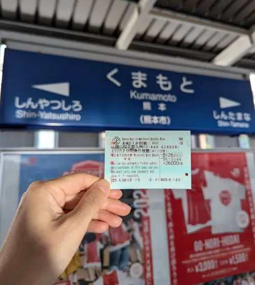 Jackie Szeto's hand, Life Of Doing, holds the Sanyo Sanin Northern Kyushu Japan Railways Pass in front of the Kumamoto Station sign