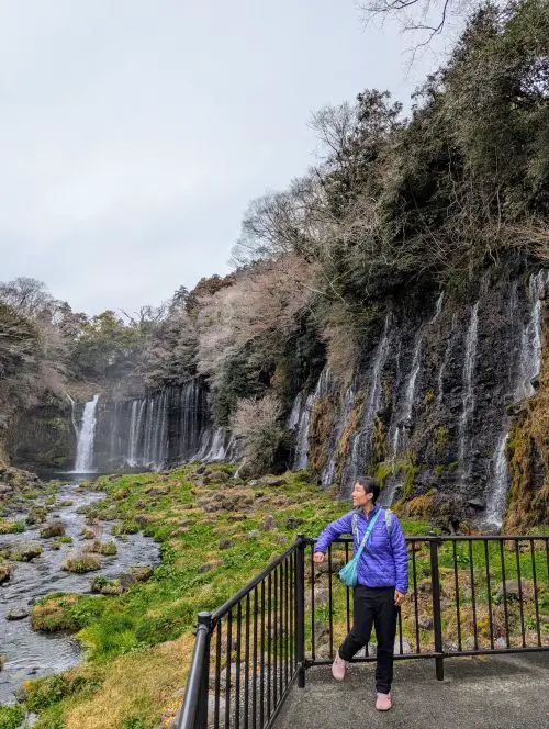 Jackie Szeto, Life Of Doing, stands next to a railing with Fujinomiya's Shiraito Waterfall in the background