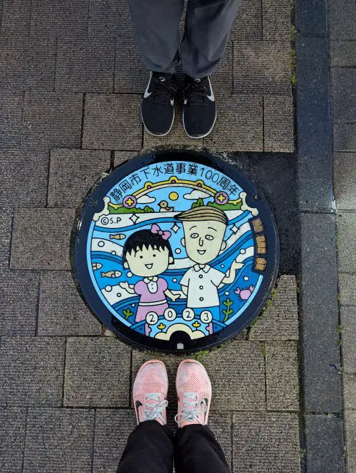 Jackie Szeto and Justin Huynh, Life Of Doing, pose with their feet next to the Chibi Maruko Chan and friend manhole cover in Shizuoka, Japan