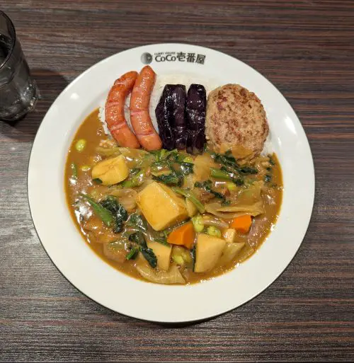 A bowl of Japanese curry rice with potato, carrots, and spring vegetables and topped with two sausages, roasted eggplant, and a hamburg patty at Shizuoka's Coco Curry Ichibanya