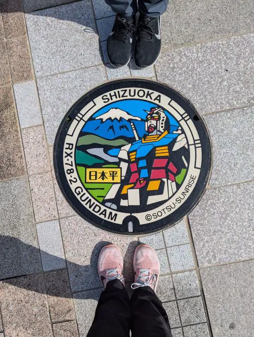 Jackie Szeto and Justin Huynh, Life Of Doing, stand around the colorful RX-78-2 Gundam manhole cover in Shizuoka, Japan