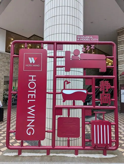 A cranberry colored art installation of a hotel themed model kit including a luggage, bath tub, and bed, located in front of the former Hotel Wing in Shizuoka, Japan