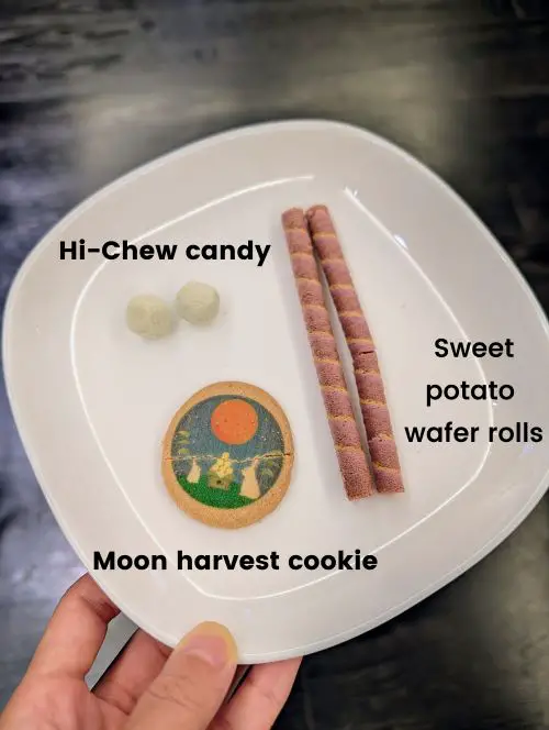 A hand holding a plate of 3 types of Japanese snacks - Hi Chew Candy, sweet potato wafer rolls, and moon harvest cookie